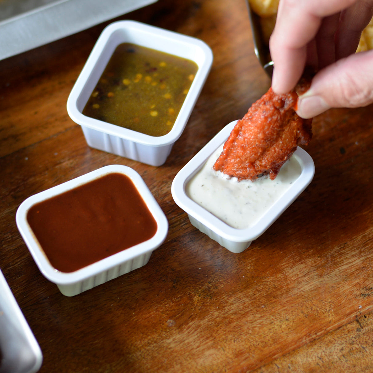 A hand dips a saucy chicken wing into ranch held in a melamine sauce cup. Nearby, two more rectangular sauce cups from the Better Burger Collection hold barbecue and vinaigrette sauces on a wooden table.
