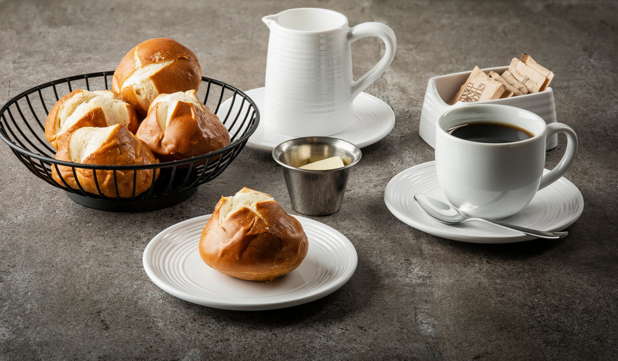 A breakfast scene with a cup of black coffee, a plate with a pretzel roll, a basket of more pretzel rolls, and a 3 oz stainless steel sauce cup filled with butter, plus a white milk pitcher and sugar packets container on a gray surface.