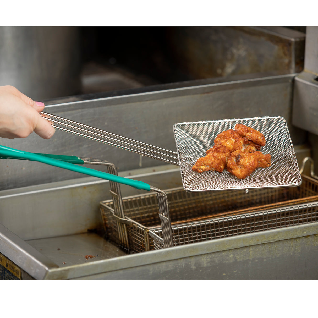 A hand holds nickel plated tongs, lifting a fine mesh wire basket filled with golden, crispy fried chicken wings out of a commercial deep fryer. The stainless steel equipment gleams in the bustling commercial kitchen background.