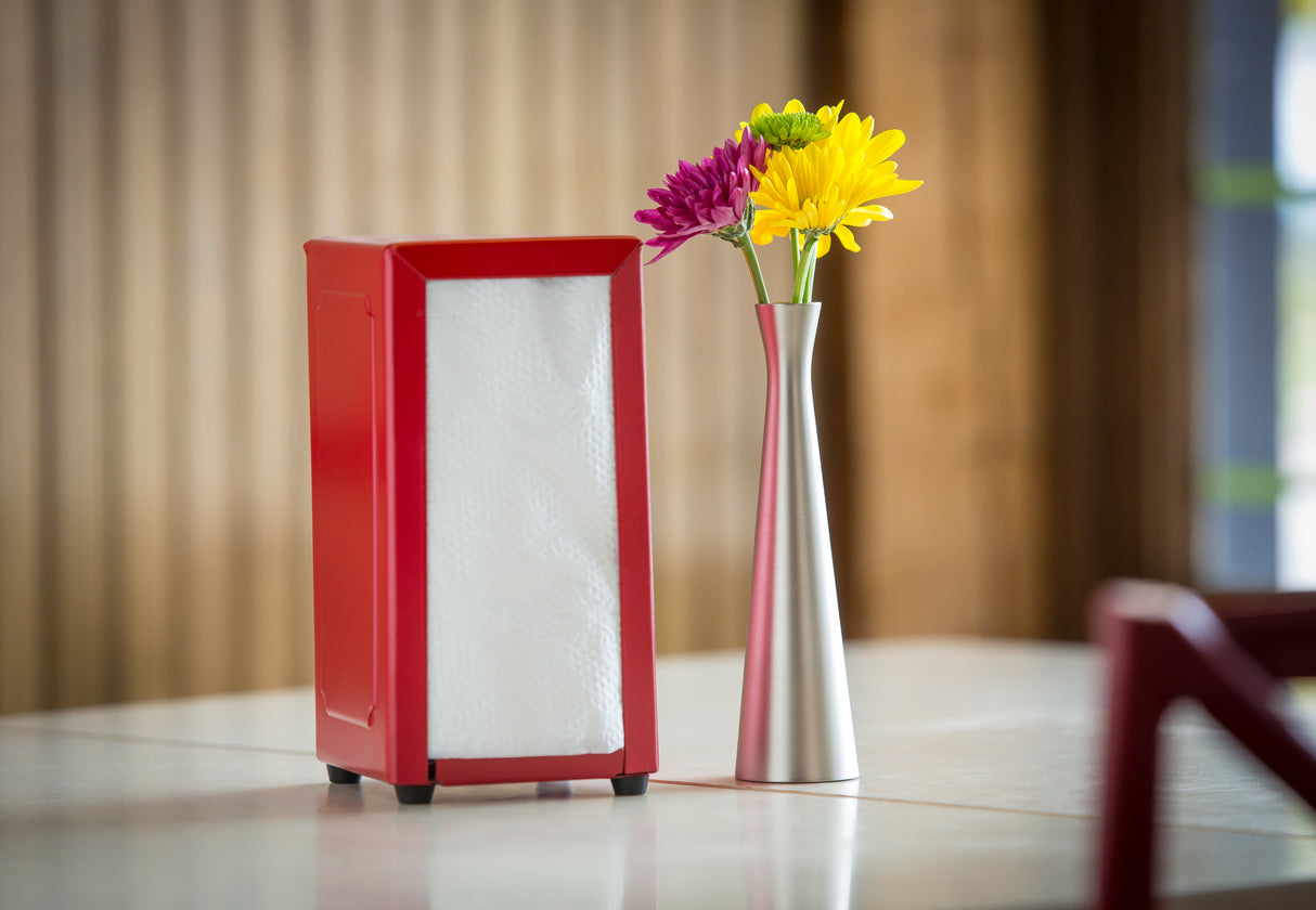 A red napkin holder with white napkins stands on a table next to a sleek zinc alloy Flower Vase with a brushed finish, holding three colorful flowers—yellow, purple, and magenta—against a softly blurred background.