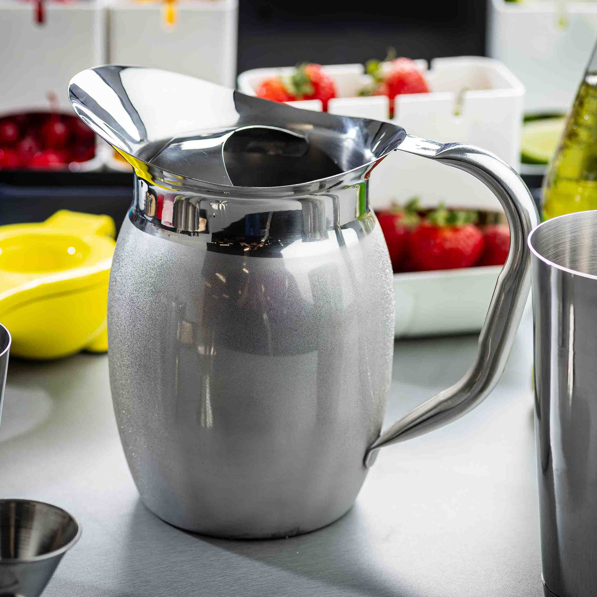 A shiny Bell Water Pitcher with a sleek handle and wide spout sits on the counter. In the background are trays of fresh fruit, a yellow citrus squeezer, and a metal cup, all suggesting a cocktail or drink prep scene.