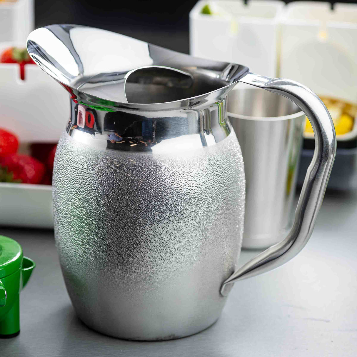 A shiny, textured Bell Water Pitcher with a wide spout and handle sits on a gray counter. In the background are blurred containers with assorted fruits, including strawberries and yellow pieces, along with a tall metal cup.