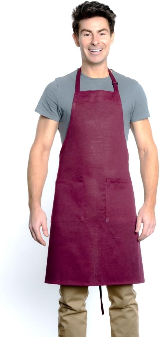 A smiling man with short dark hair wears a gray t-shirt, tan pants, and a dark red bib apron with an adjustable neck and two front pockets. He stands facing forward against a plain white background, arms relaxed at his sides.