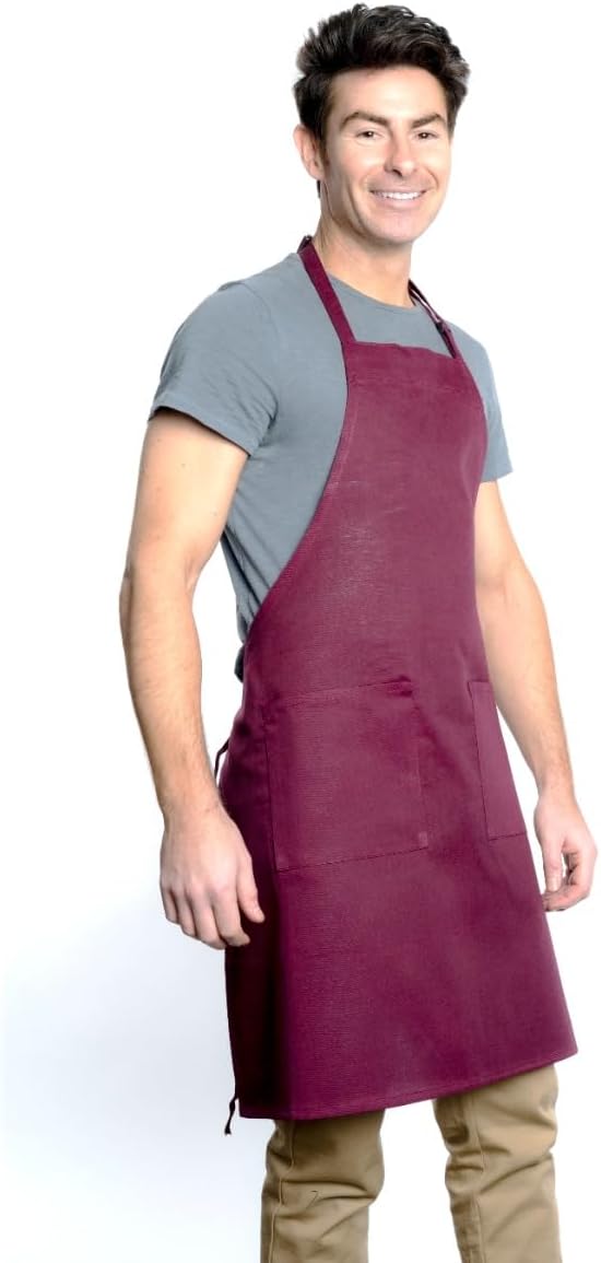 A smiling man with short dark hair wears a gray t-shirt, khaki pants, and a chef approved maroon bib apron with an adjustable neck and front pockets, standing against a plain white background.