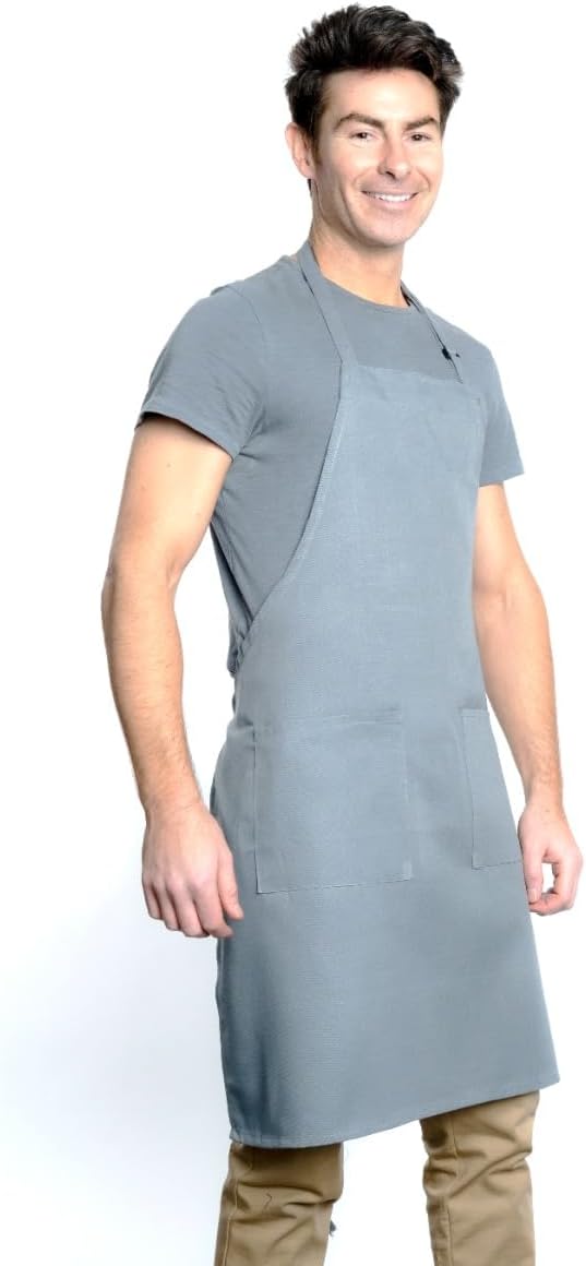 A smiling man with short brown hair wears a chef approved light gray bib apron with front pockets and an adjustable neck, over a light gray T-shirt and tan pants. He stands against a white background, facing slightly to the right with relaxed arms.