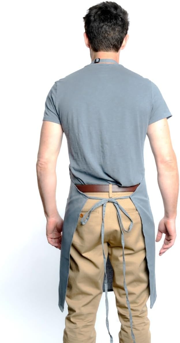 A man stands facing away from the camera, wearing a gray short-sleeve shirt, tan pants, and a chef-approved bib apron with an adjustable neck. The apron’s straps are visible, and his arms are relaxed at his sides against a plain white background.