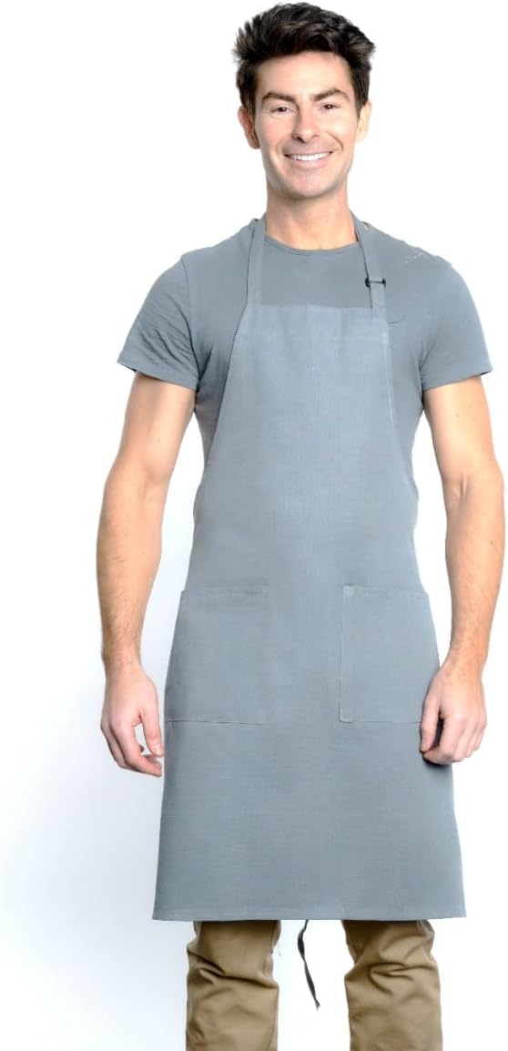 A smiling man with short dark hair wears a plain gray T-shirt, tan pants, and a chef approved bib apron with an adjustable neck and two front pockets. He stands facing forward against a white background, hands relaxed at his sides.