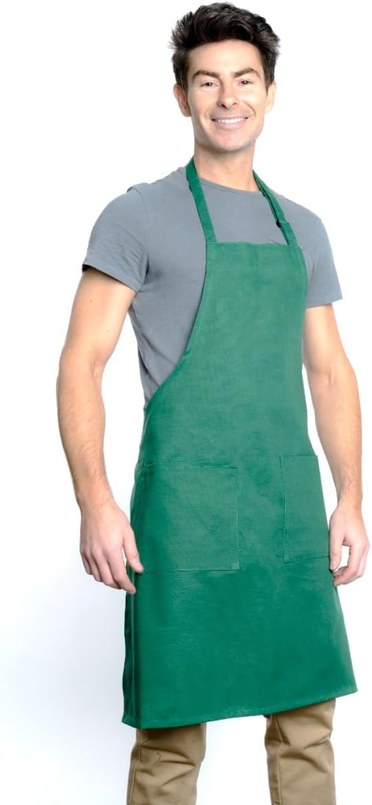 A smiling man with short dark hair wears a gray T-shirt, tan pants, and a chef-approved green bib apron with two front pockets and an adjustable neck. He stands against a plain white background, facing forward, left arm slightly bent.
