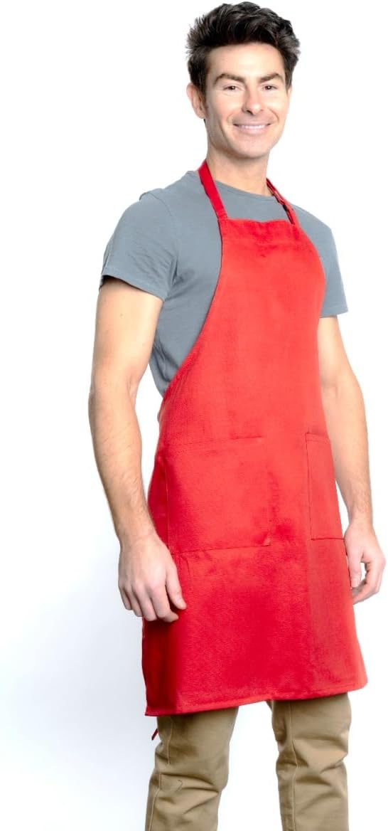 A smiling man with short dark hair wears a gray t-shirt, tan pants, and a bright red bib apron with an adjustable neck and front pockets. He stands in a relaxed pose against a plain white background.