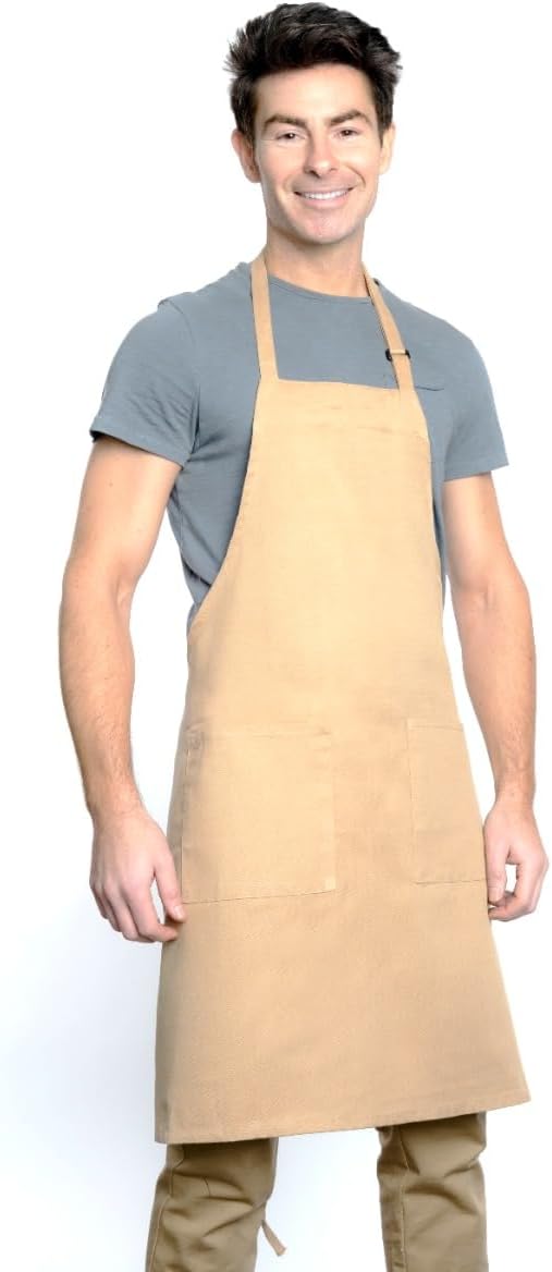A man with short, dark hair smiles while wearing a chef approved bib apron featuring an adjustable neck, over a gray t-shirt and tan pants. The apron has two front pockets and a neck strap, set against a plain white background.