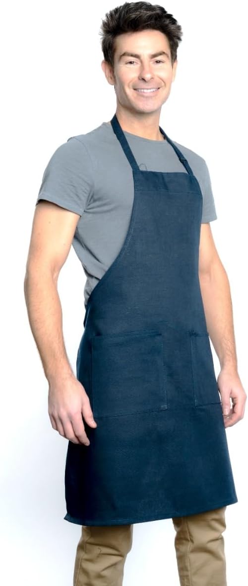 A smiling man with short dark hair wears a gray T-shirt, tan pants, and a chef approved bib apron with an adjustable neck and two front pockets. He stands against a plain white background, facing slightly to the left with one hand resting by his side.