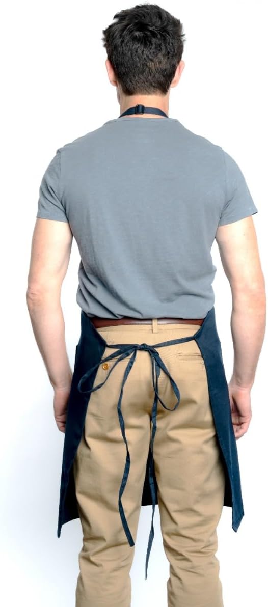 A man stands facing away, wearing a gray T-shirt, tan pants, and a chef approved dark blue bib apron with an adjustable neck, tied at the waist and neck. His arms are relaxed at his sides against a plain white background.