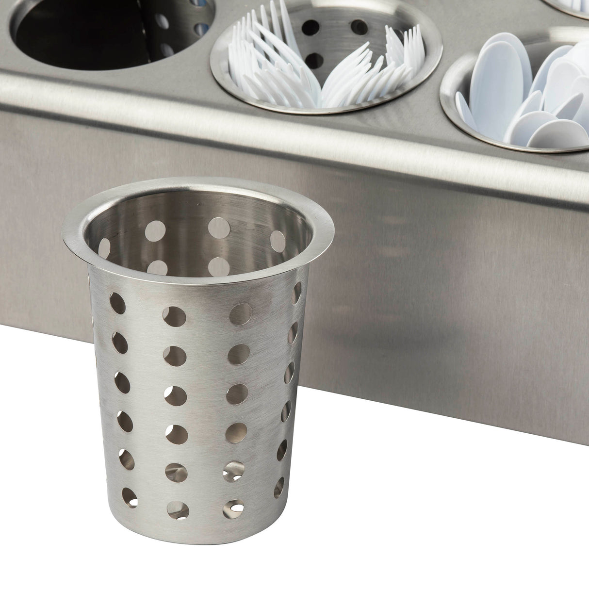 A stainless steel flatware cylinder with round holes sits in front of a metal countertop organizer filled with similar, dishwasher safe holders containing white plastic spoons and forks—ideal for organized kitchen or food service storage.