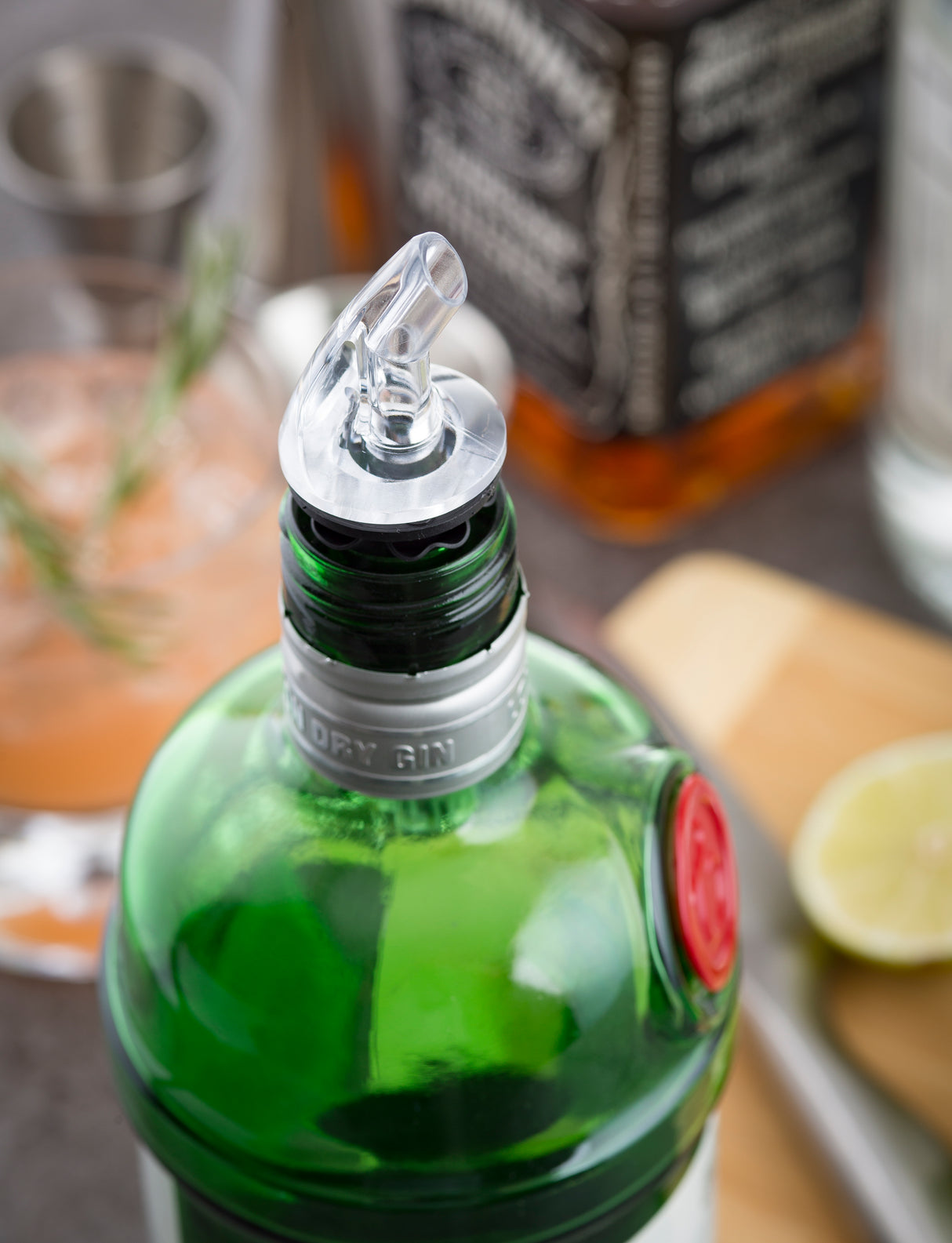 A close-up of a green gin bottle with a BPA free Proper Pour Widemouth Pourer attached. In the blurred background, there’s a cocktail with rosemary, sliced lime, a jigger, and another premium liquor bottle, suggesting a drink-making scene.