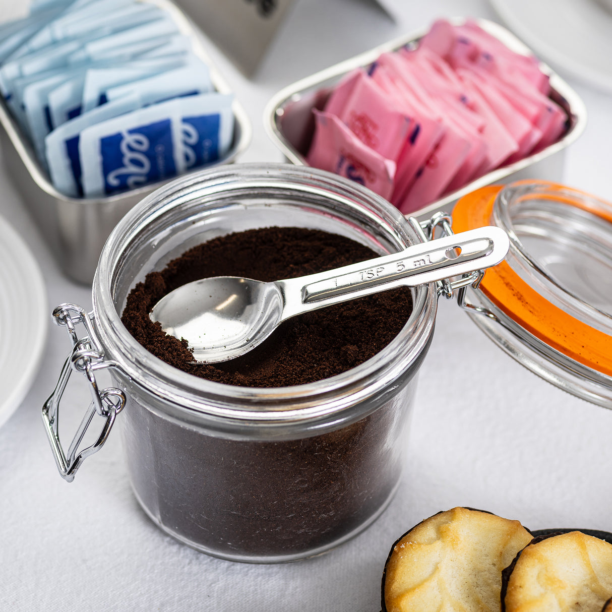 An open glass jar filled with ground coffee sits on a white surface, with a stainless steel teaspoon inside. Nearby, metal containers hold blue and pink sweetener packets, and part of a pastry is visible in the foreground.