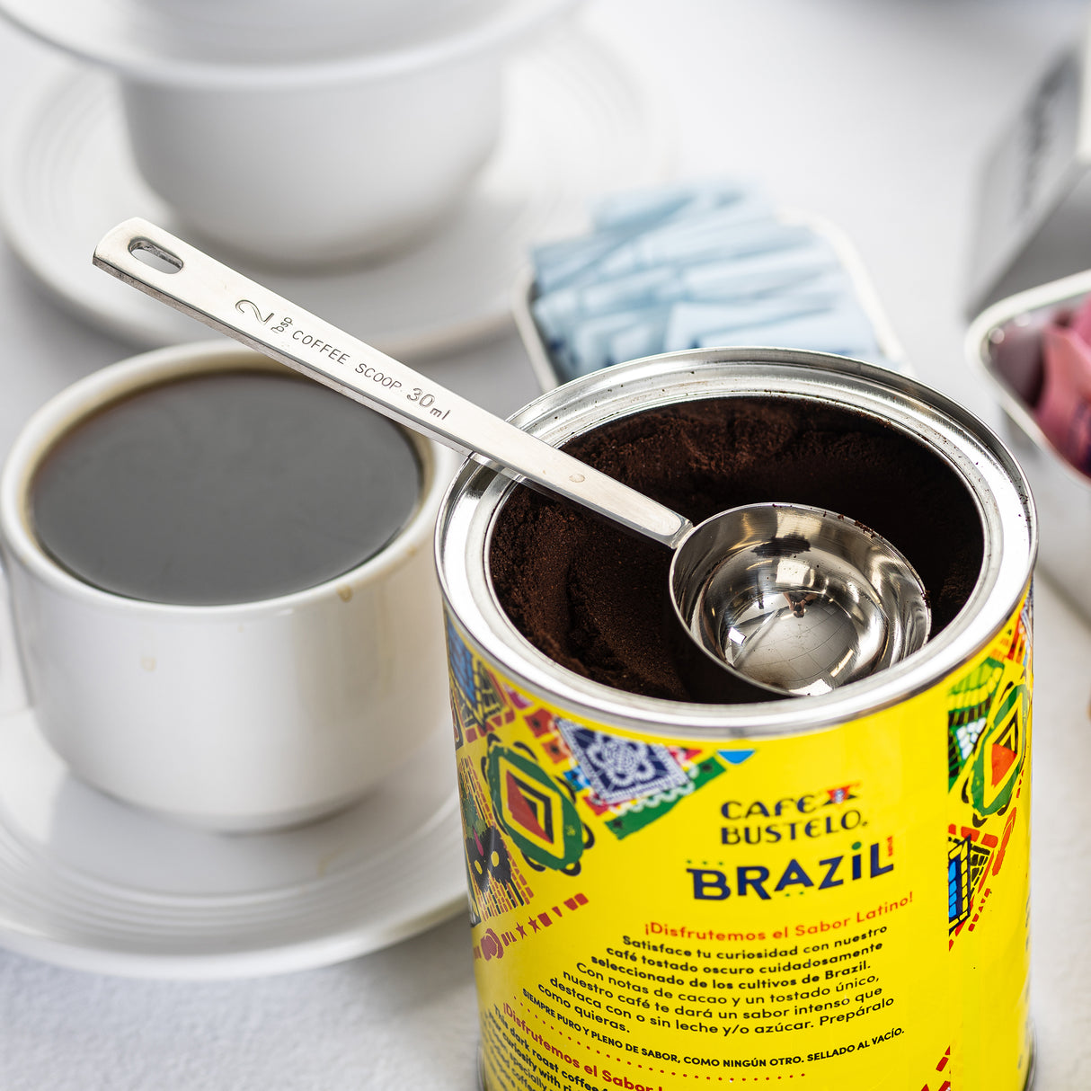 An opened can of Café Bustelo Brazil coffee with a stainless steel coffee scoop inside is shown. Next to it is a cup of black coffee in a white cup and saucer, and a container holding blue and pink sweetener packets on a white surface.