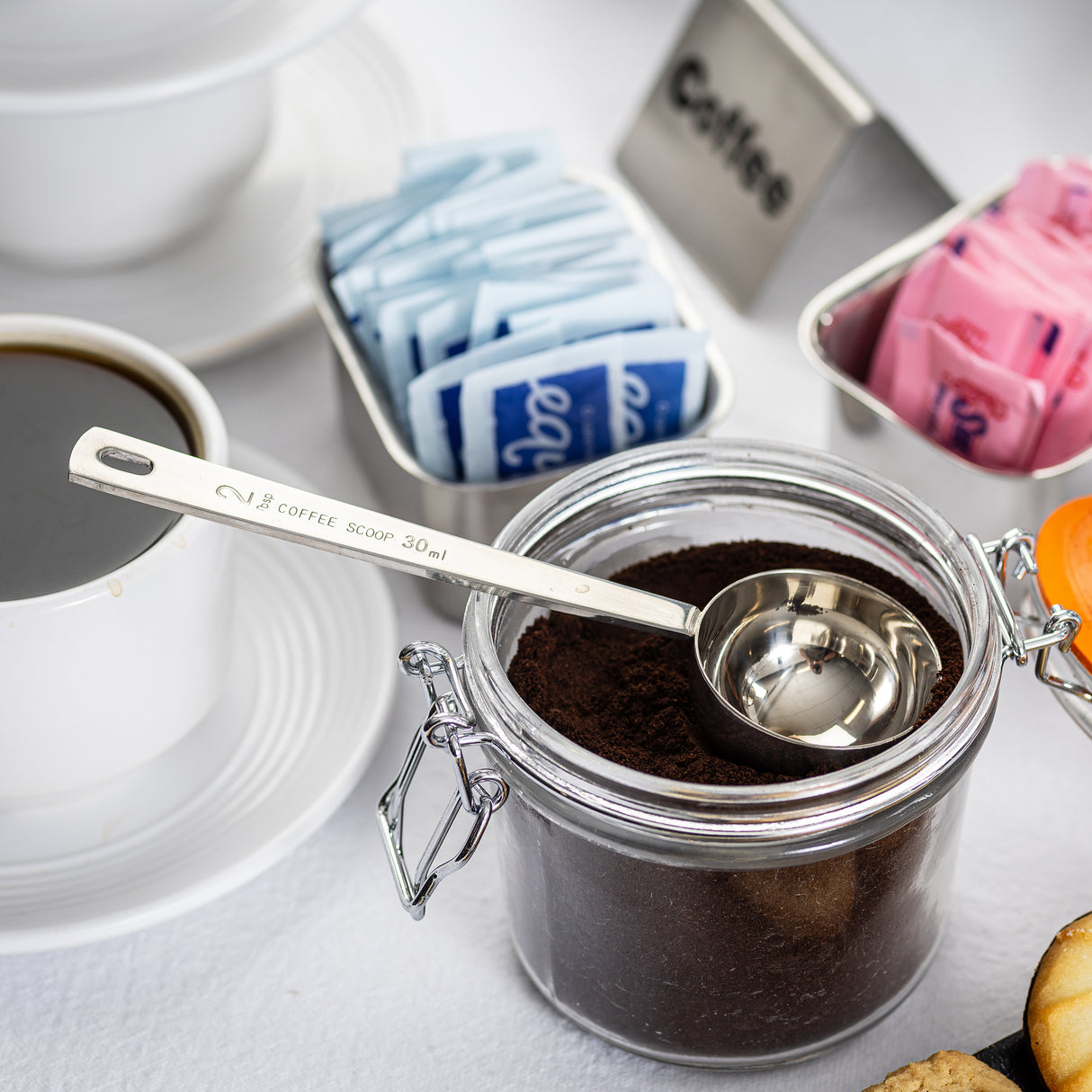 A glass jar of ground coffee with a stainless steel 2 tablespoon Coffee Scoop inside sits on a table. Nearby are a cup of black coffee, blue and pink sweetener packets in metal containers, and part of a plate with pastries on a white tablecloth.