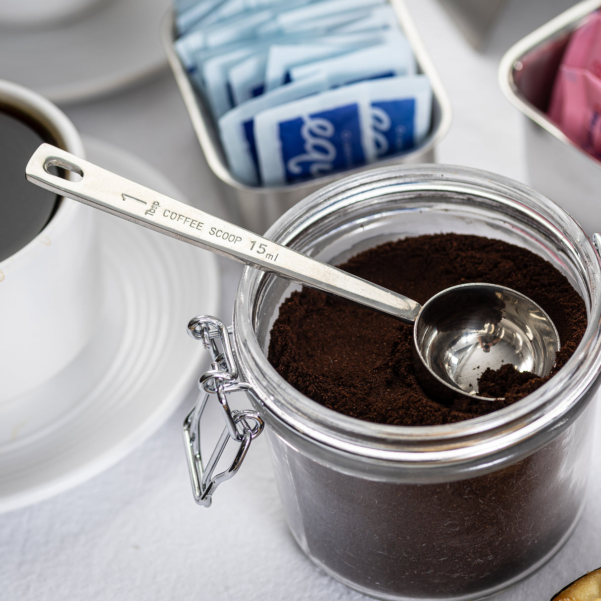 A long handled stainless steel measuring spoon rests in a glass jar filled with ground coffee. Nearby are a cup of coffee and a metal tray with blue and pink sweetener packets on a white surface. The scoop is labeled COFFEE SCOOP 15 ml.