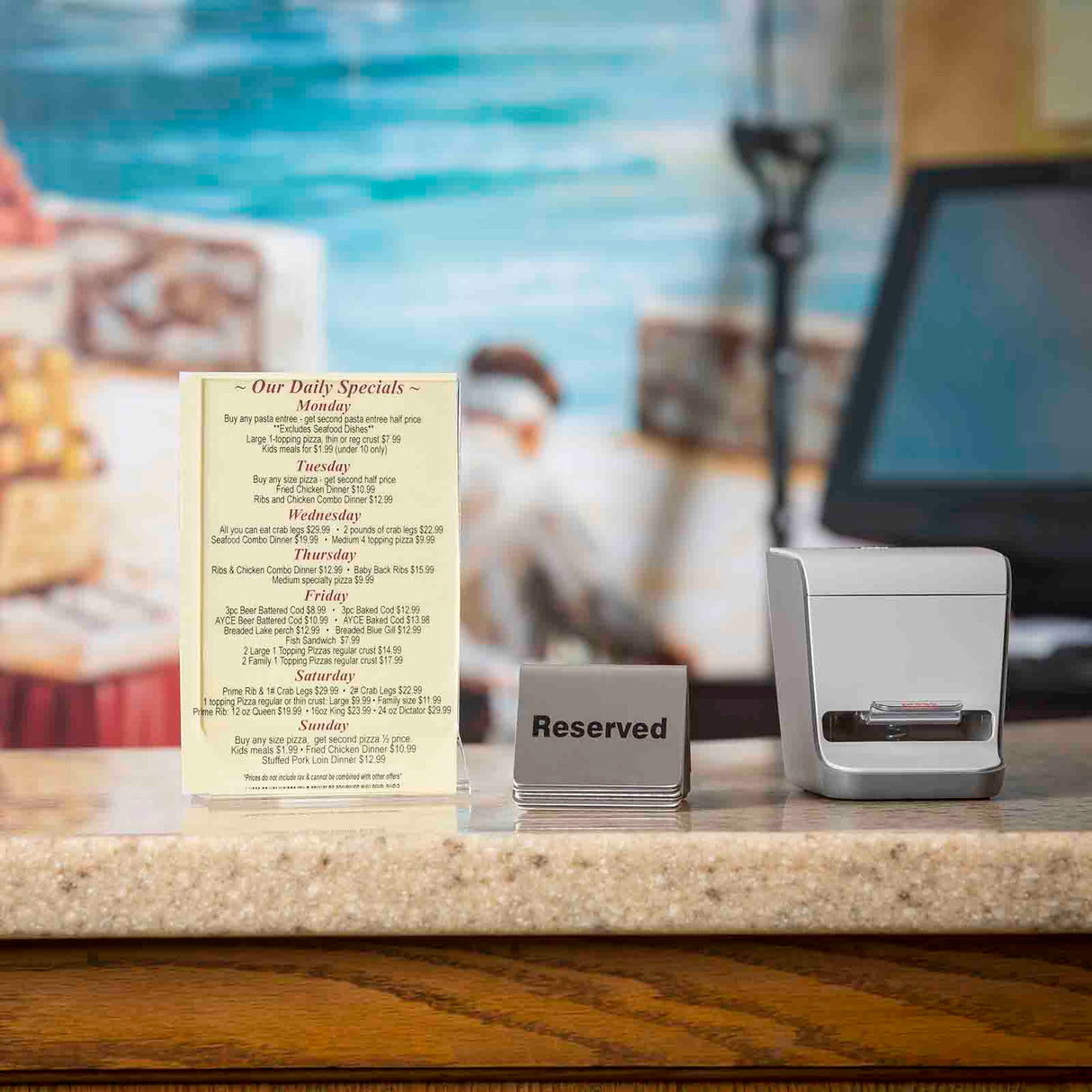 A restaurant counter with a daily specials menu in an acrylic menu holder, a Reserved sign, and a receipt printer. The blurred background features a mural with a figure by the sea. The beige countertop highlights the signs and printer up front.