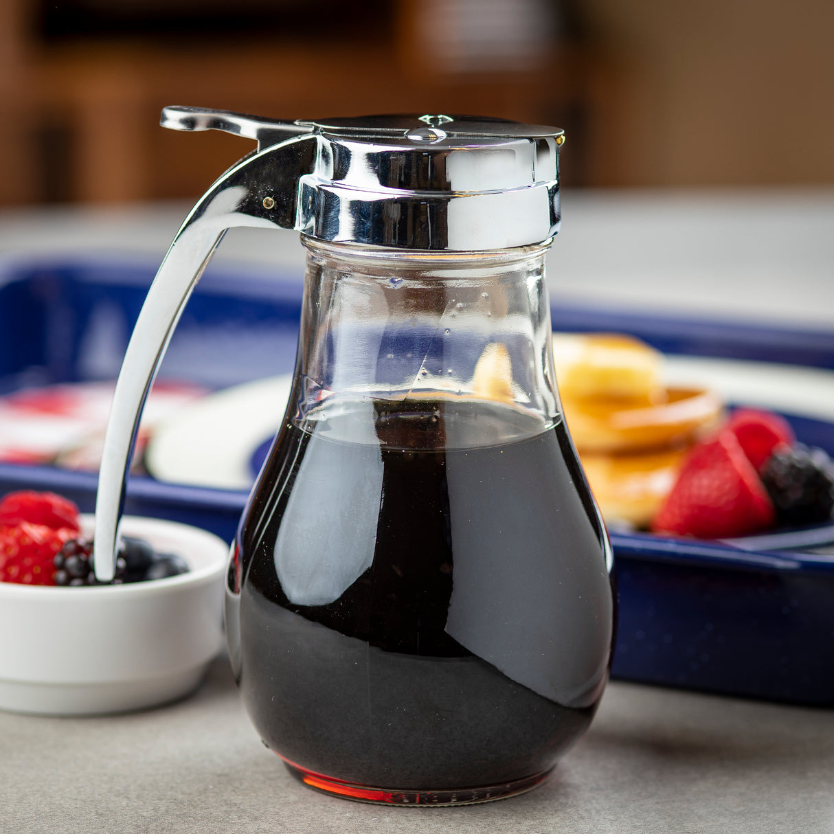A clear TableCraft 414 glass syrup dispenser with a shiny metal lid and handle, filled with dark syrup, sits on a countertop. In the blurred background, berries and pancakes are partially visible, hinting at a cozy breakfast setting.
