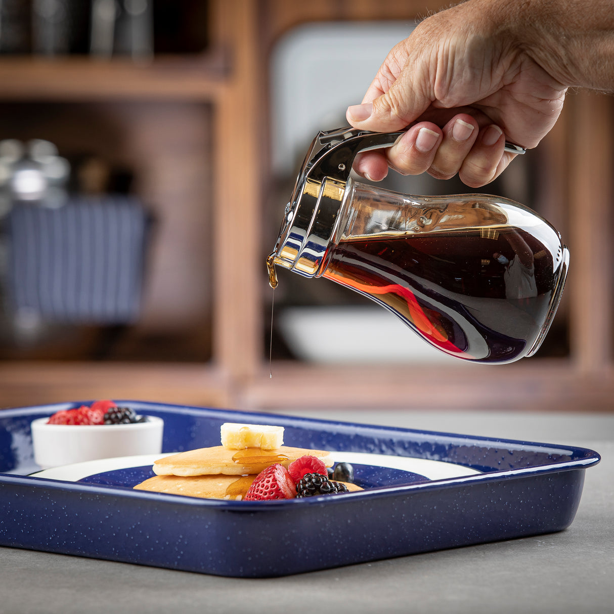 A hand pours syrup from a TableCraft 414 glass pitcher onto pancakes topped with butter. The pancakes are on a blue plate with strawberries, blackberries, and a small white bowl of more berries. The pitcher is dishwasher safe.