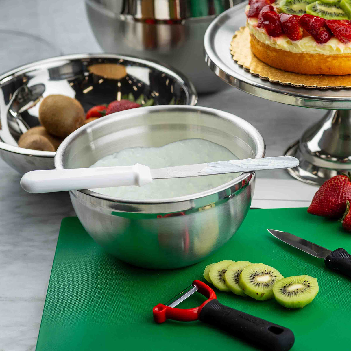 A kitchen scene with a green cutting board holding sliced kiwi, a peeler with a stainless steel blade, and a paring knife. A metal bowl of whipped cream and an icing spatula rest nearby, with whole kiwis, strawberries, and a fruit-topped cake in the background.
