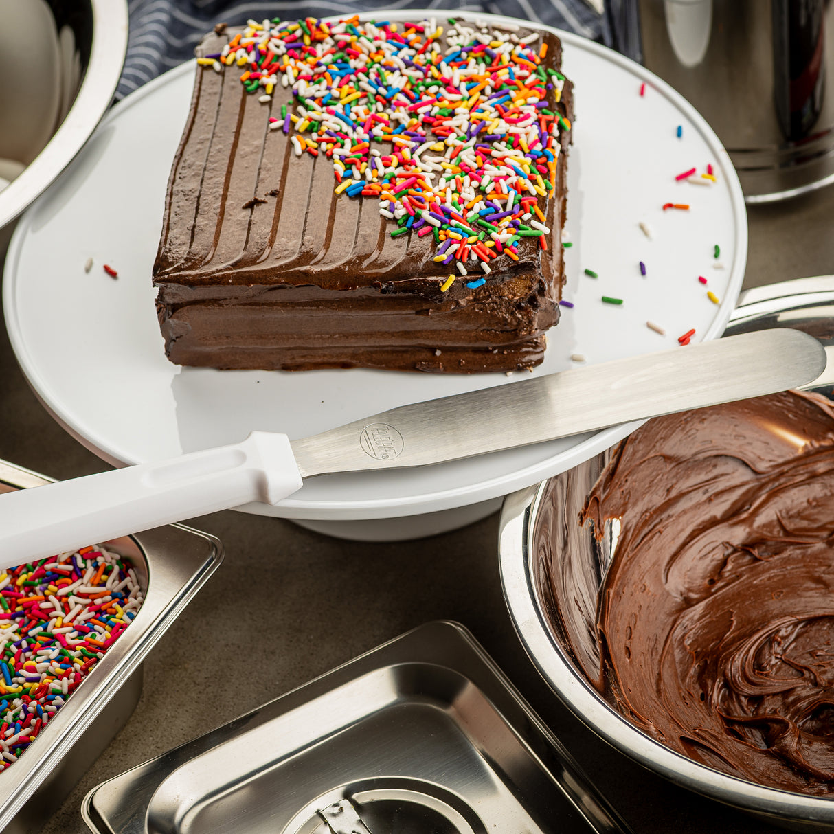 A square chocolate cake with chocolate frosting and rainbow sprinkles on one corner sits on a white plate. An icing spatula with a stainless steel blade and white handle rests nearby, above a bowl of chocolate frosting. Metal trays with more sprinkles surround the cake.