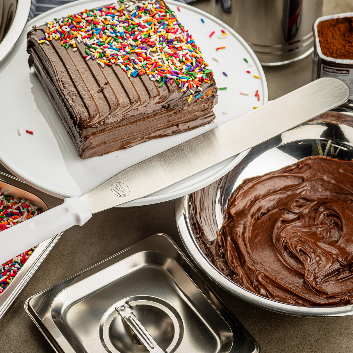 A square chocolate cake with chocolate frosting and rainbow sprinkles sits on a white plate. An icing spatula with a stainless steel blade rests nearby, surrounded by bowls of frosting, sprinkles, and baking tools on the countertop.