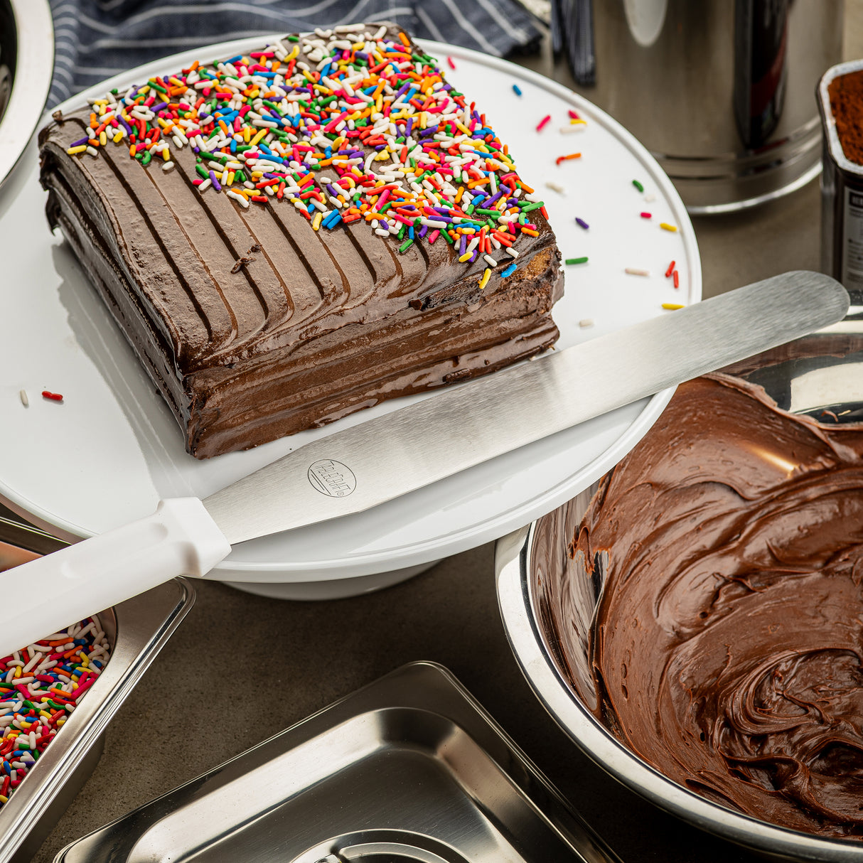 A rectangular chocolate cake with chocolate frosting and rainbow sprinkles sits on a white cake stand. A large icing spatula with a stainless steel blade rests on the stand. Nearby, a metal bowl holds chocolate frosting and a tray is partially in view.