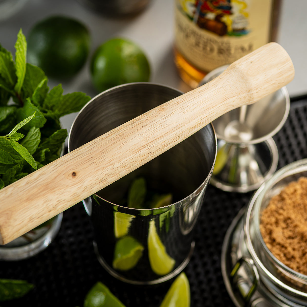 A natural wood TableCraft muddler rests on a metal shaker containing lime wedges. Fresh mint, whole limes, a jar of brown sugar, a jigger, and rum are arranged nearby, suggesting the makings of a classic mojito cocktail.