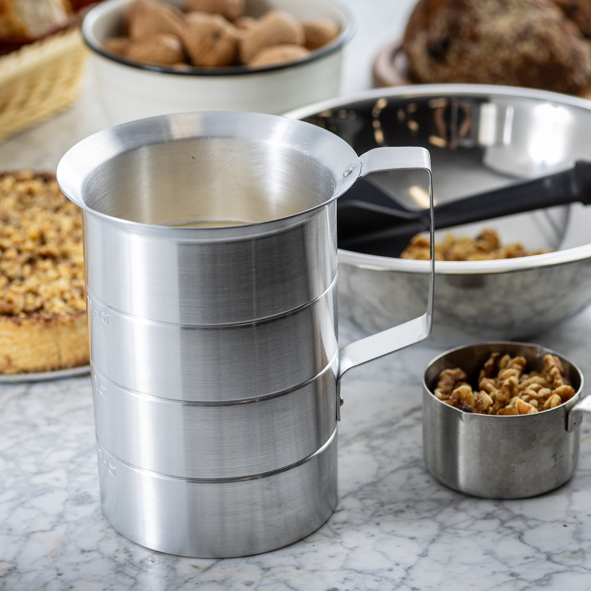 A large aluminum flour sifter with a handle sits on a marble countertop, surrounded by baking ingredients including walnuts in a graduated measure, a walnut cake, a bowl of nuts, and a metal bowl with utensils in the background.