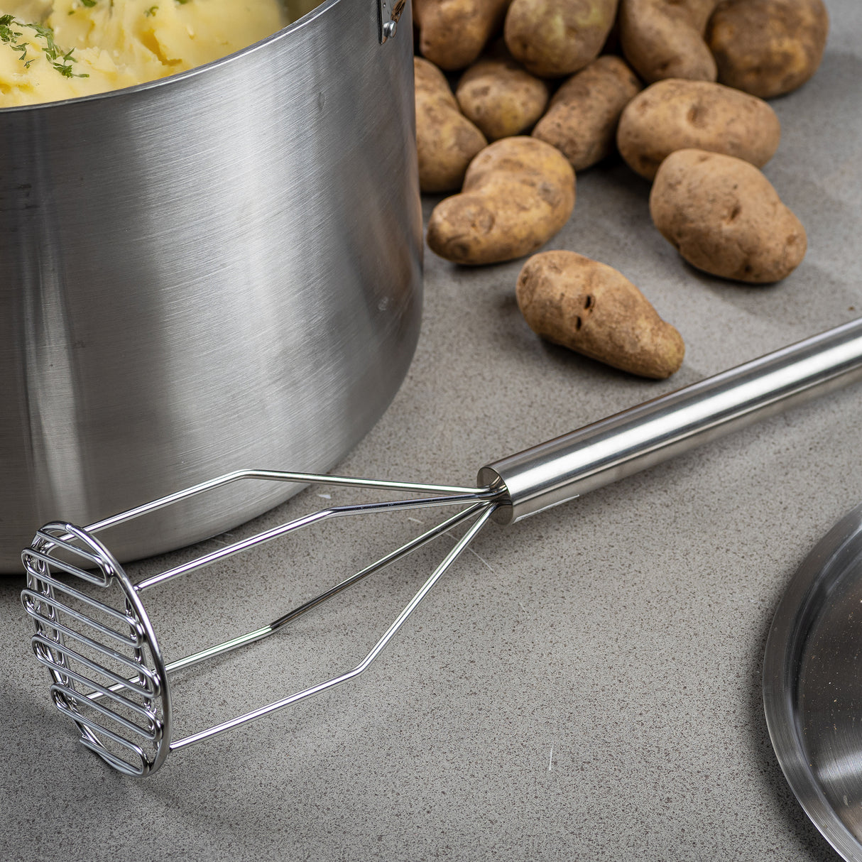 A Potato Masher with a stainless steel handle rests on a gray countertop beside a large pot of mashed potatoes, whole unpeeled potatoes, and a pot lid. The masher is dishwasher safe for easy cleanup.