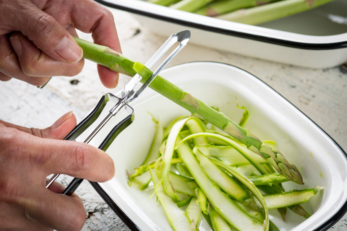 A close-up of hands using a peeler with a stainless steel blade to shave thin strips from a green asparagus stalk over a white enamel dish. Peeled asparagus strips are collected in the dish, with more stalks visible in the background.