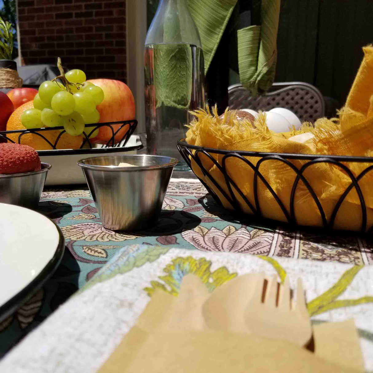 A close-up of a table set outdoors with a plate, wooden cutlery, and napkin in the foreground. A stainless steel sauce cup sits beside a basket of yellow fabric with two eggs, fruit bowl, metal cup, and glass bottle. Green leaves and brick wall behind.