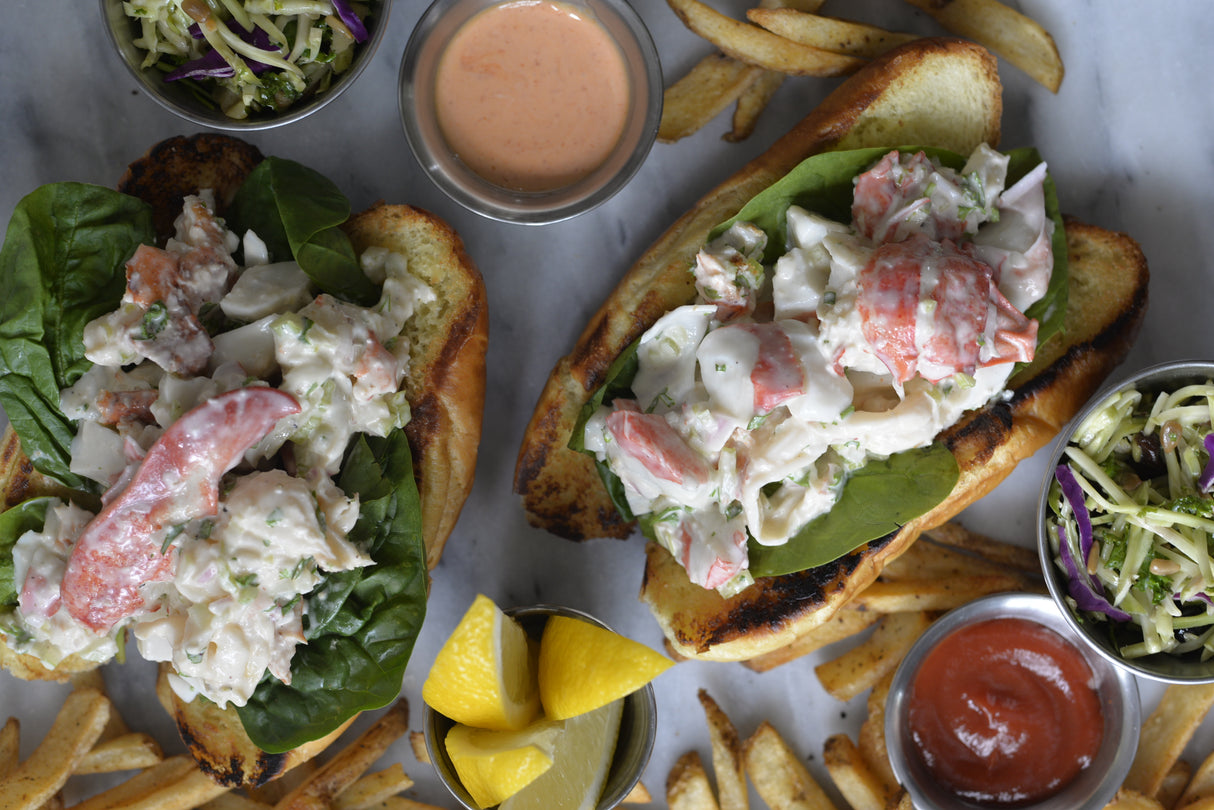 Two toasted lobster rolls filled with lobster salad and greens sit on a white surface, surrounded by golden fries, lemon wedges, coleslaw, and dipping sauces served in sleek stainless steel sauce cups. The scene is vibrant and appetizing with fresh, colorful ingredients.