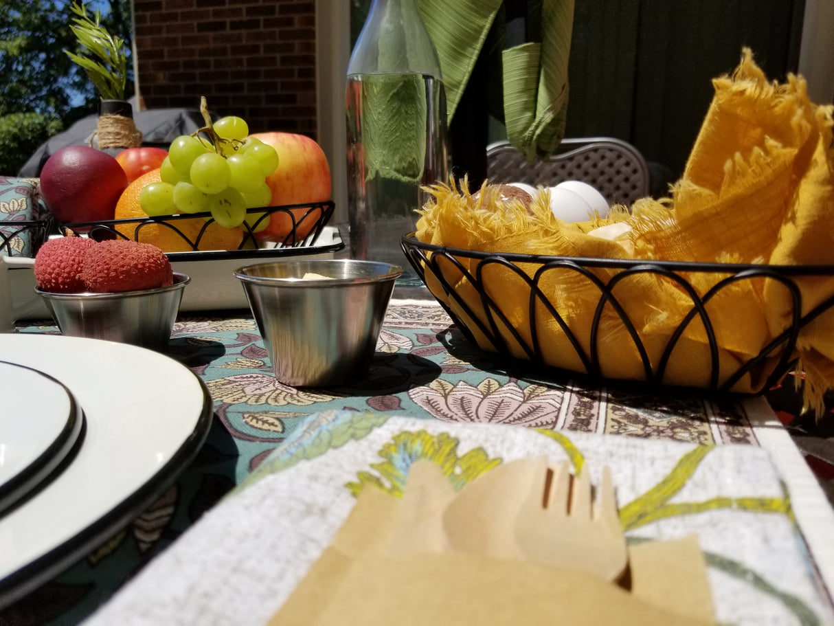 A close-up of an outdoor table set for a meal, featuring a basket of yellow napkins, plates, stainless steel cutlery, bowls of fruit including apples, grapes, lychees, a glass bottle, and a sauce cup on a floral tablecloth in bright sunlight.