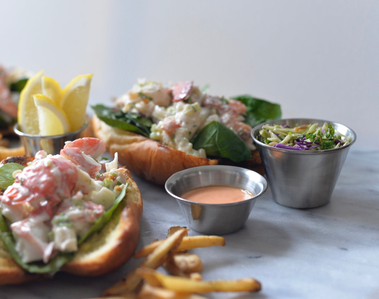 A lobster roll with chunks of lobster, spinach, and creamy dressing sits on a toasted bun. Sides include lemon wedges, thin fries, a 6 oz stainless steel sauce cup, and coleslaw with green and purple cabbage on a marble surface.
