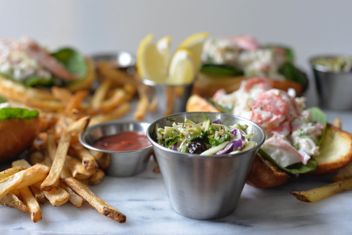 A close-up of a meal featuring a stainless steel, dishwasher safe sauce cup of coleslaw in the foreground, with thin French fries, ketchup, lemon wedges, and lobster rolls on toasted buns arranged on a marble surface.
