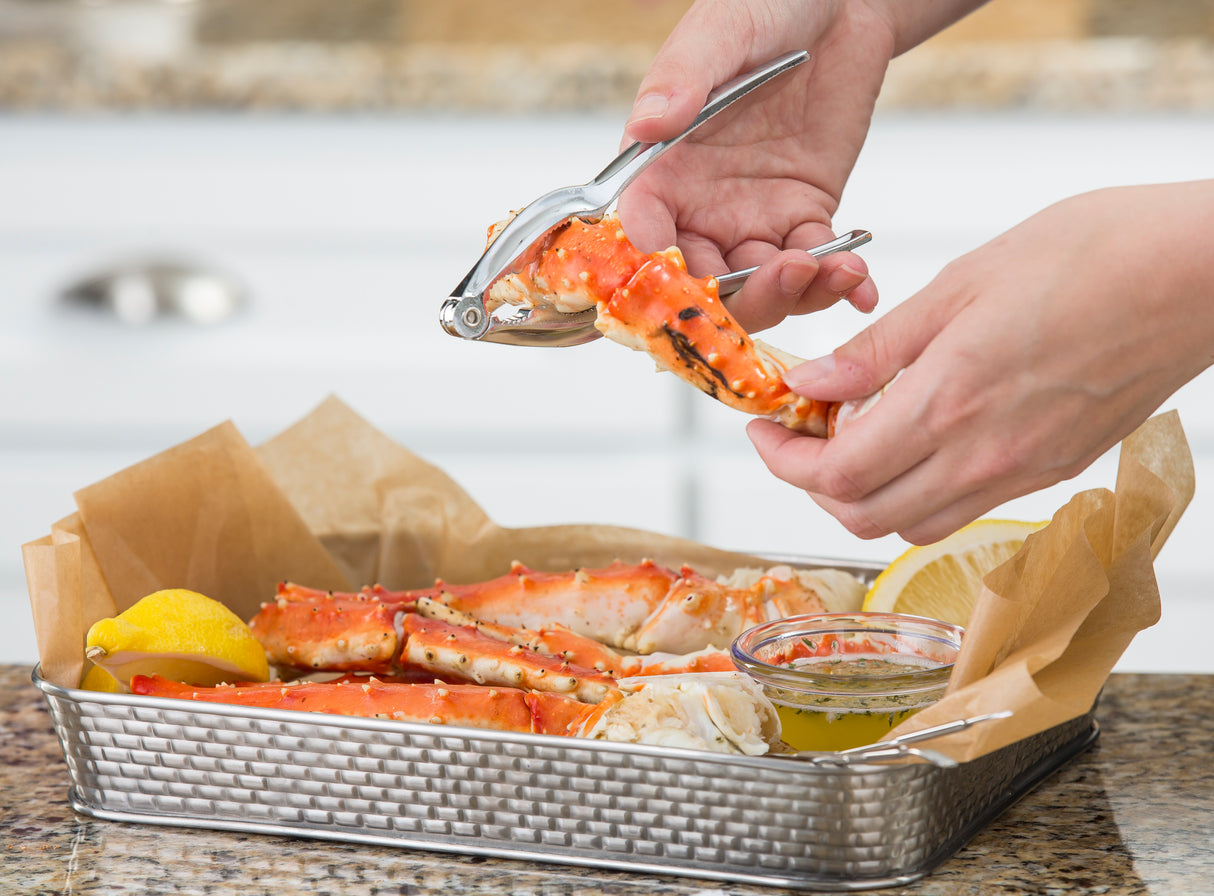 A person uses a chrome plated seafood cracker to open a red and white crab leg over a basket lined with parchment paper. Inside the basket are more crab legs, lemon wedges, and a glass dish of butter sauce on a granite counter.