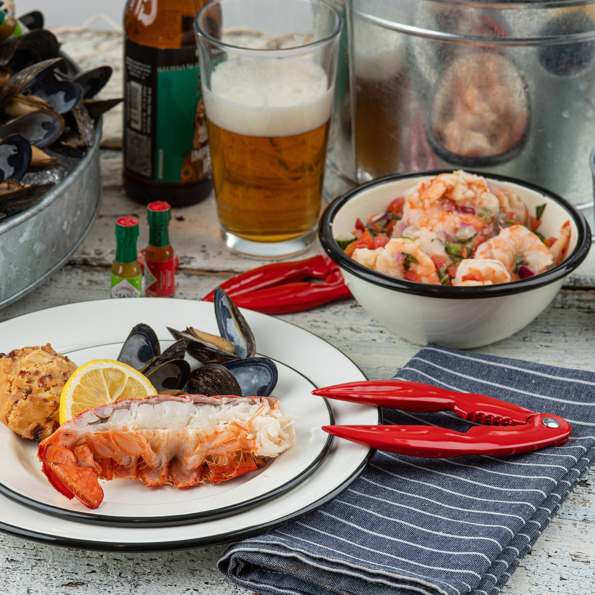 A plate with lobster tail, mussels, a lemon slice, and a biscuit sits on a striped napkin. Nearby are red enamel shell crackers, beer, shrimp salad in a bowl, hot sauce bottles, and buckets of mussels and shrimp on a rustic wooden table.