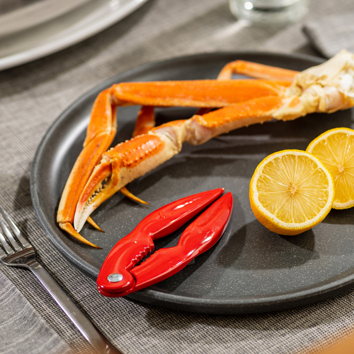 A black plate holds a crab leg, a red enamel cast aluminum cracker, and two lemon halves, all arranged on a gray placemat next to a fork. The scene suggests preparation for enjoying seafood with the perfect lobster crack tool.