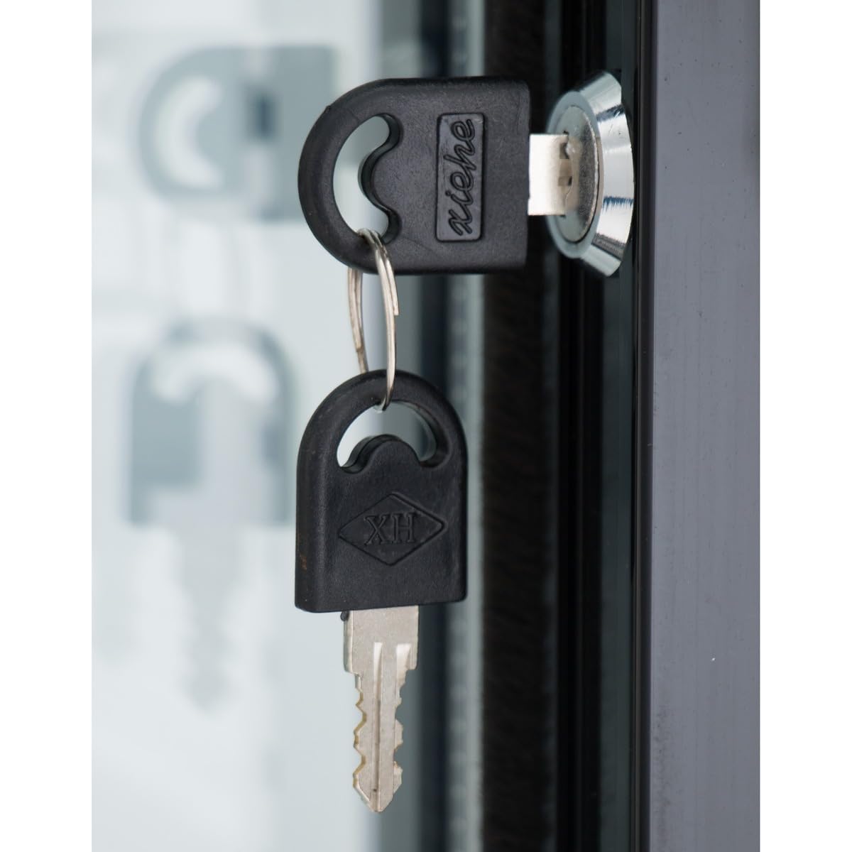 A close-up of a key inserted into the lock of a commercial refrigerator, with a second identical key hanging from the keyring. The keys have black plastic heads with logo engravings; the metallic frame reflects light in the background.