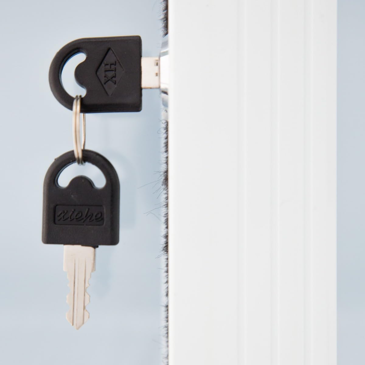 Close-up of two silver keys with black plastic heads in a white door lock, ideal for Empura Refrigeration units or refrigerated merchandisers with glass sliding doors. One key is inserted, while the other hangs below on a metal ring. Soft blue background.