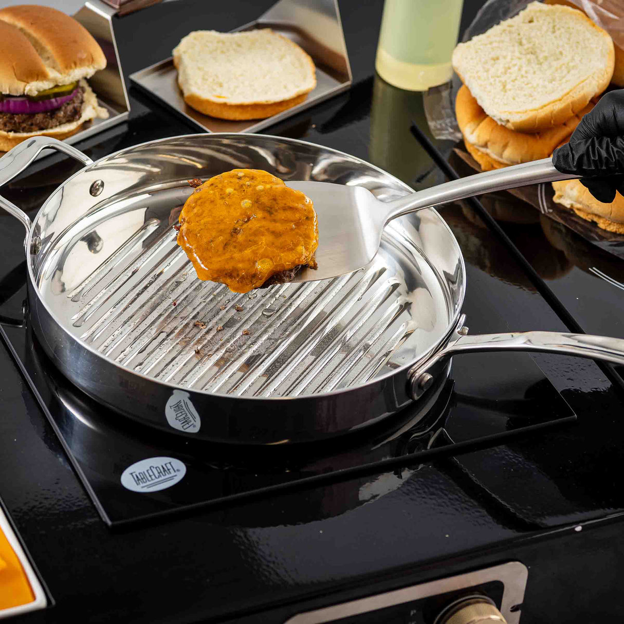A hand in a black glove uses a Dalton Collection Turner to lift a cooked burger patty with melted cheese from a stainless steel grill pan. Open buns and toppings like lettuce, onion, and tomato are arranged nearby, ready for assembling.