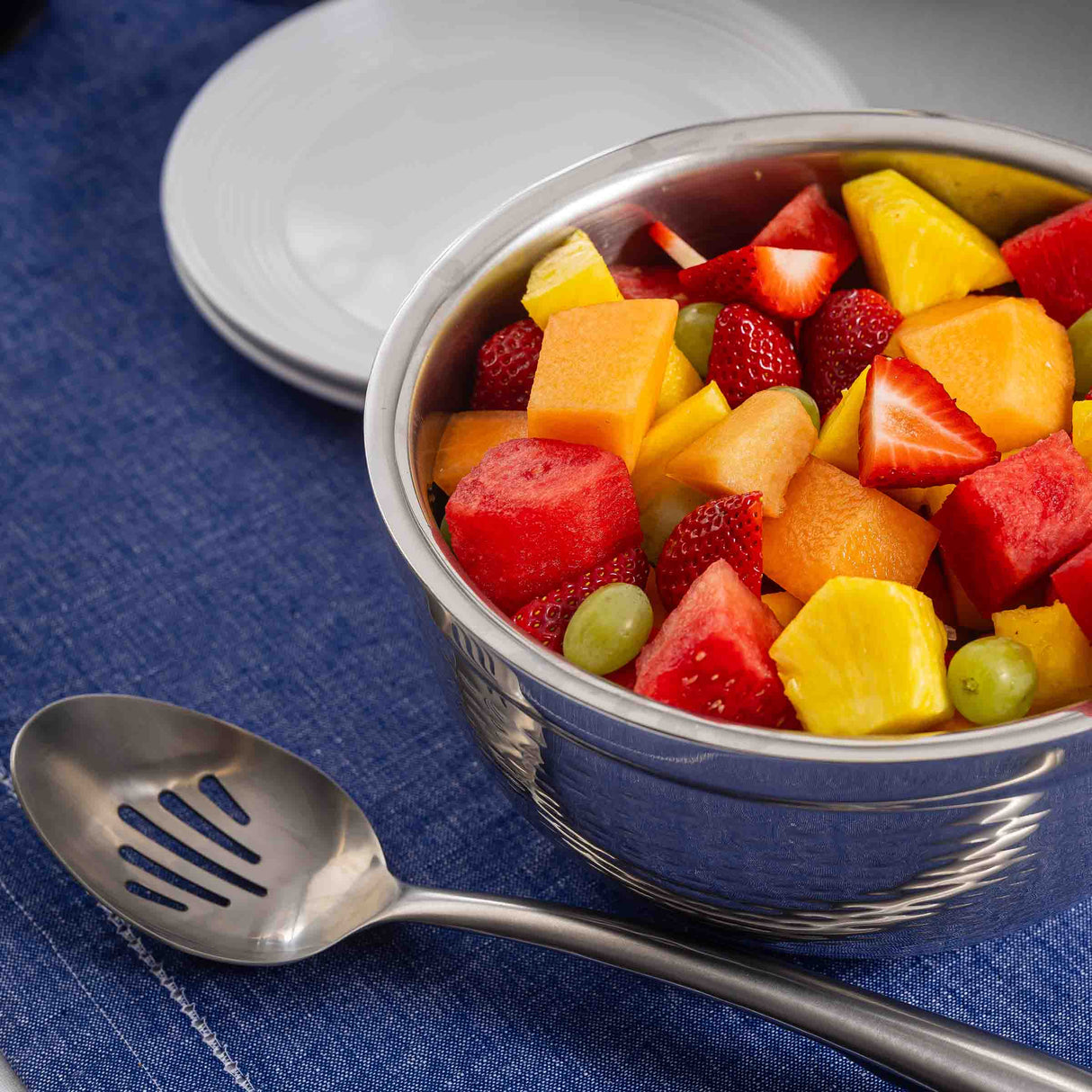 A stainless steel bowl filled with mixed fresh fruit—watermelon, cantaloupe, pineapple, strawberries, and green grapes—sits on a blue cloth. A Dalton Collection Buffet Spoon and white plates are nearby, ready for serving.