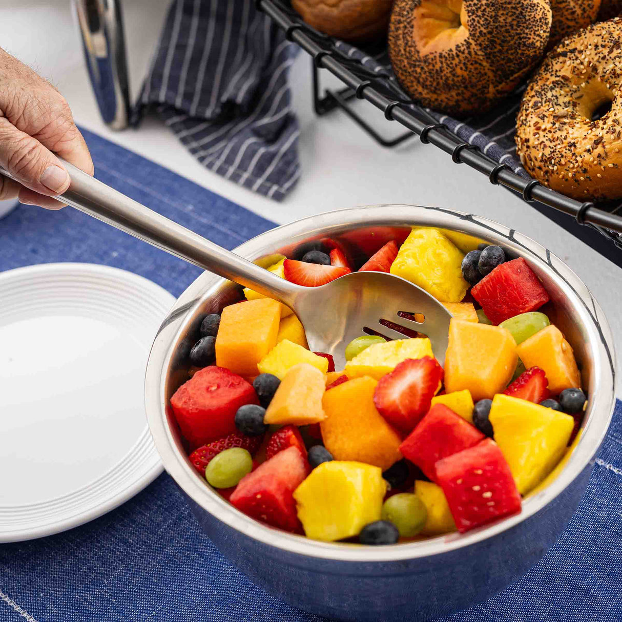 A hand holds a Dalton Collection Buffet Spoon over a metal bowl filled with colorful fruit salad, including strawberries, watermelon, pineapple, and more. Bagels and a stack of white plates sit nearby on a blue cloth.