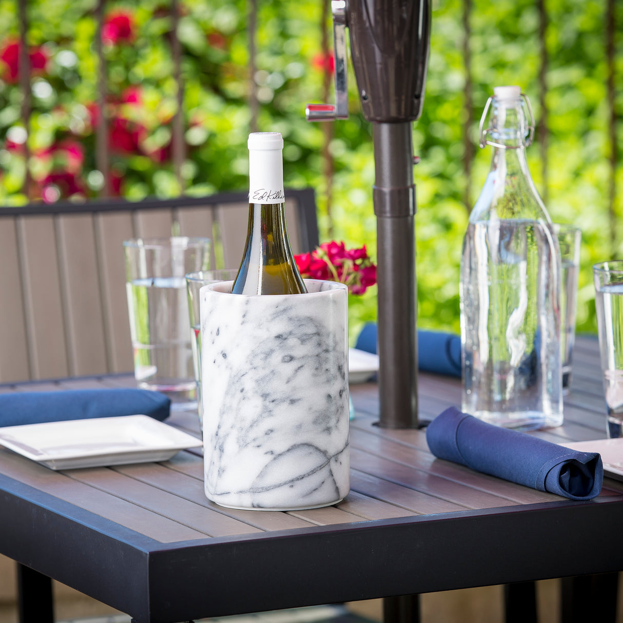 A chilled white wine bottle in a white marble wine cooler sits on an outdoor table set with glasses, water bottle, navy napkins, and white plates. Bright greenery and red flowers are visible in the blurred background. TableCraft elegance shines through.