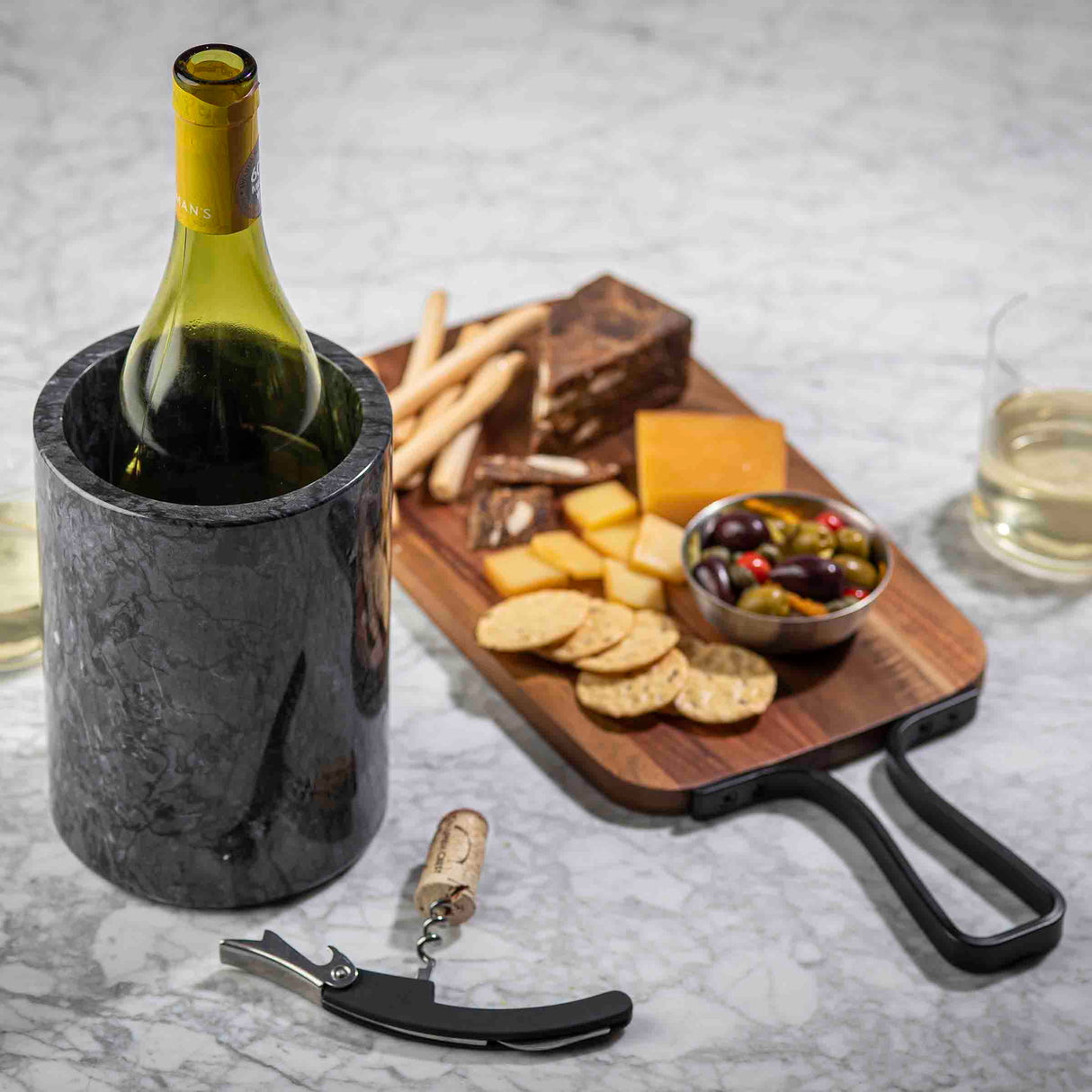 A wine bottle rests in a black marble TableCraft wine cooler beside a corkscrew on a marble table. Behind it, a wooden board offers cheese, crackers, breadsticks, meats, and olives. Two glasses of white wine complete the scene.