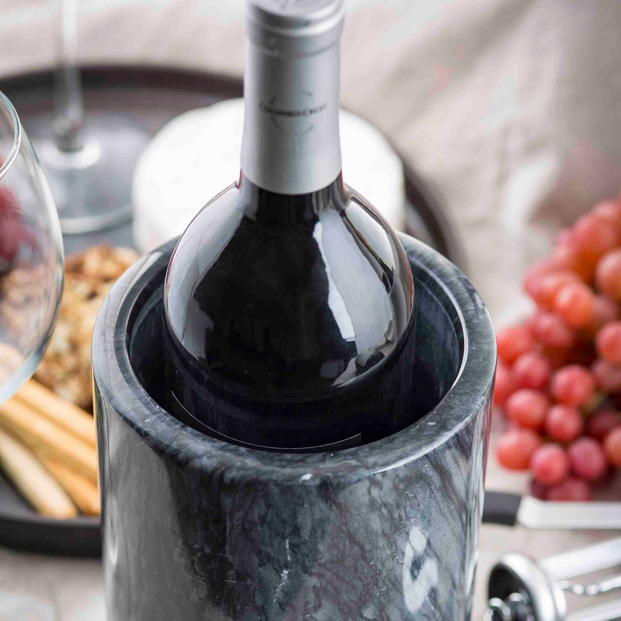 A bottle of red wine rests in a TableCraft black marble wine cooler. In the background, a tray holds grapes, breadsticks, assorted cheeses, and walnuts beside a wine glass, creating an elegant appetizer or entertaining setting.