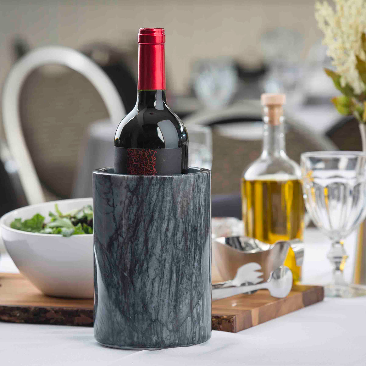 A bottle of red wine with a red foil top sits in a black marble TableCraft wine cooler on a white tablecloth. Nearby are a glass oil bottle, a glass of water, a salad bowl, and a gravy boat on a wooden board, with dining blurred in the background.